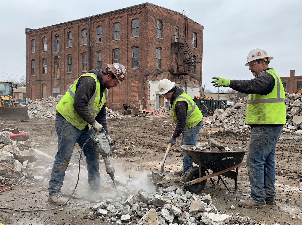 A three-person crew in hi-vis vests and hard hats breaking up concrete in front of a large brick commercial building