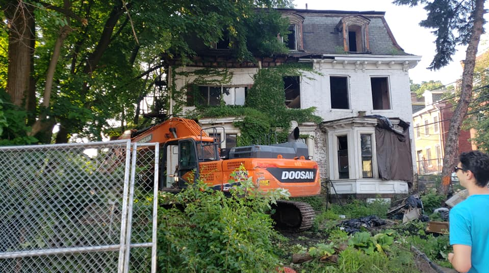 An excavator positioned in front of a large, overgrown, vine-covered Victorian house prior to demolition